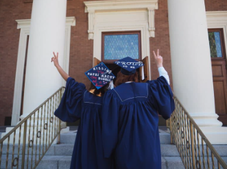 Two students in graduation cap and gown shown from the back, each flashing peace signs.
